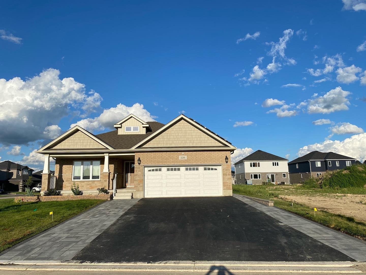 A suburban house with a two-car garage under a clear blue sky. Neighboring homes and grassy lawns are visible against a backdrop of fluffy clouds.