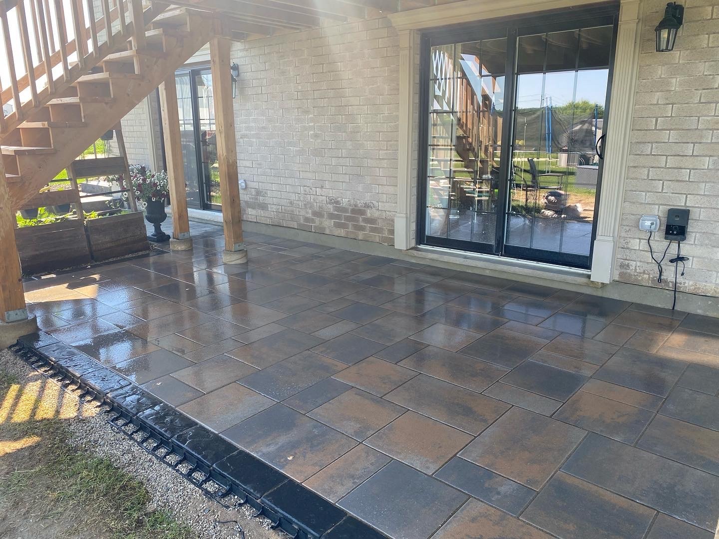 Outdoor patio with wet tile flooring under a wooden staircase. Sliding glass door reflects yard furniture and trampoline. Planters decorate the space.