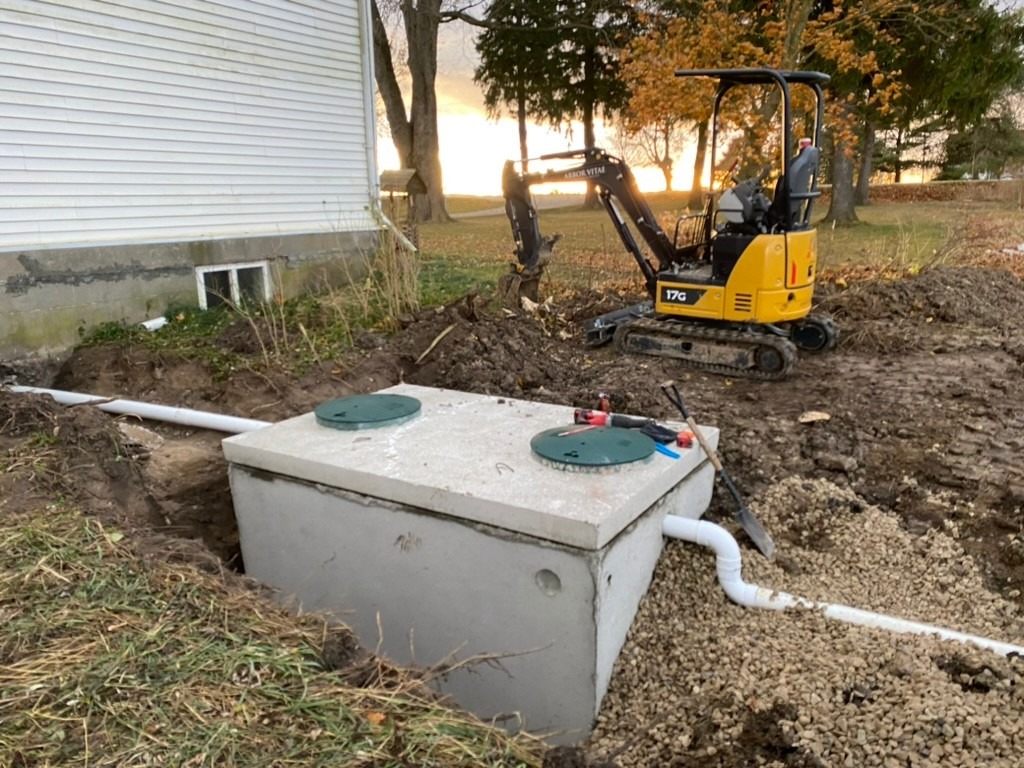 A construction site features a small excavator and a newly installed septic tank beside a white house, surrounded by trees at sunset.