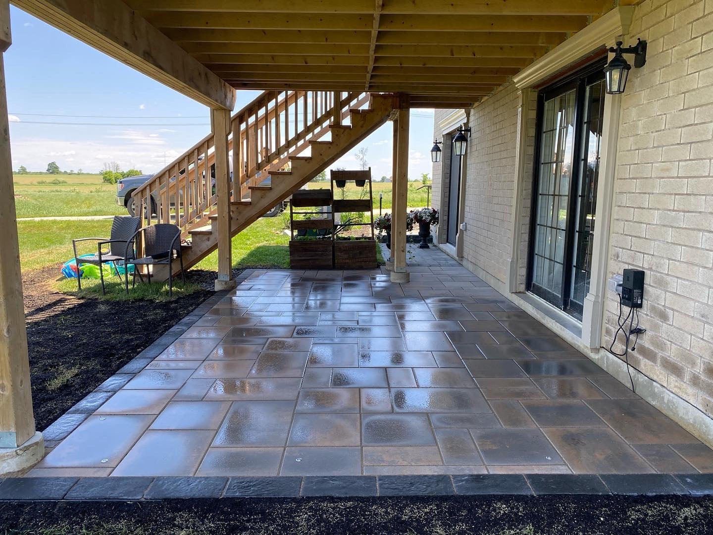 Outdoor patio with wet tiles, wooden staircase, and chairs. Vehicle and open field in background, near a house with sliding glass doors.