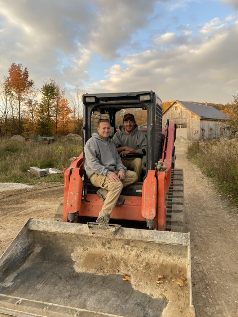 Two people sit in a small excavator on a dirt path. Autumn trees and a rustic barn are in the background.