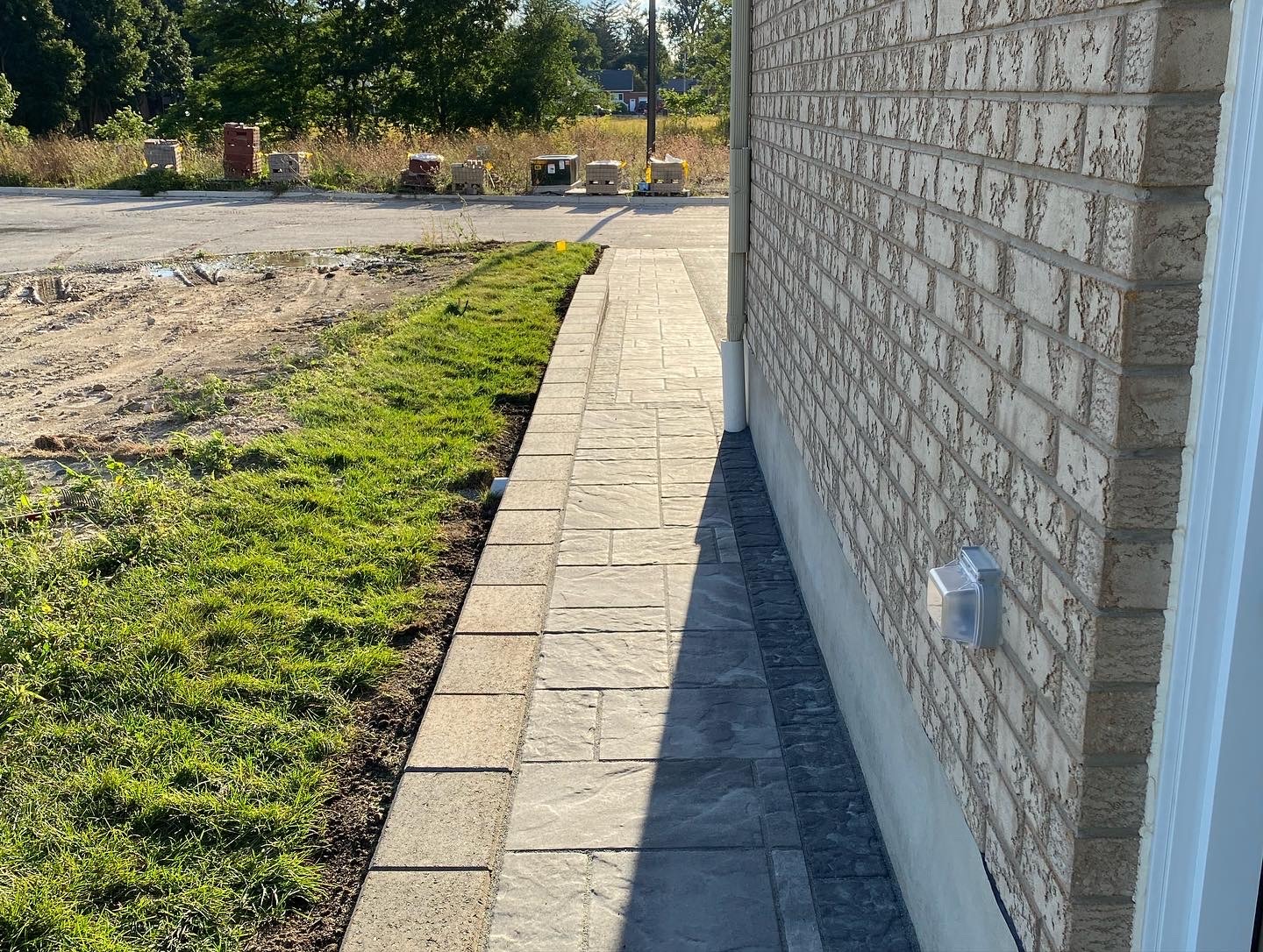 Brick building with a paved walkway and grassy edge leading to a partially cleared area with stacked materials in the background.