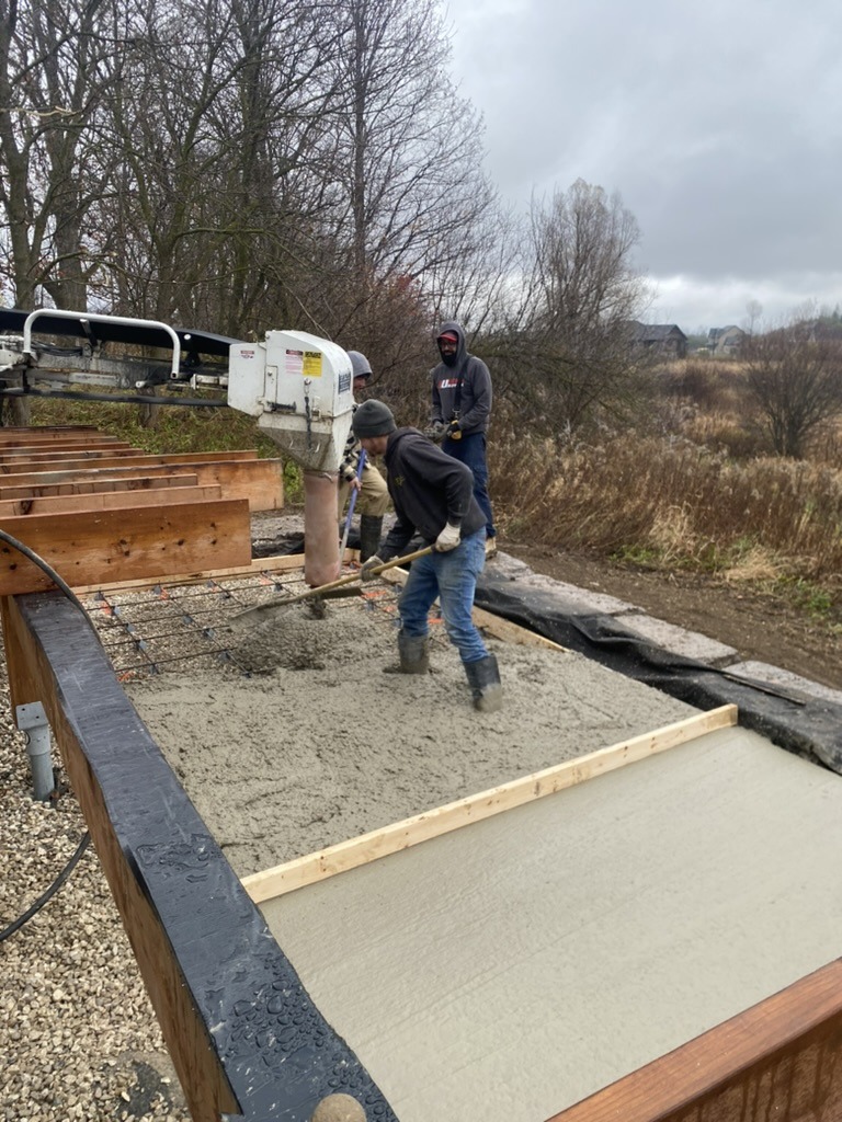 Two people are working outdoors, pouring concrete on a rectangular framework using a large machine, surrounded by bare trees and a cloudy sky.