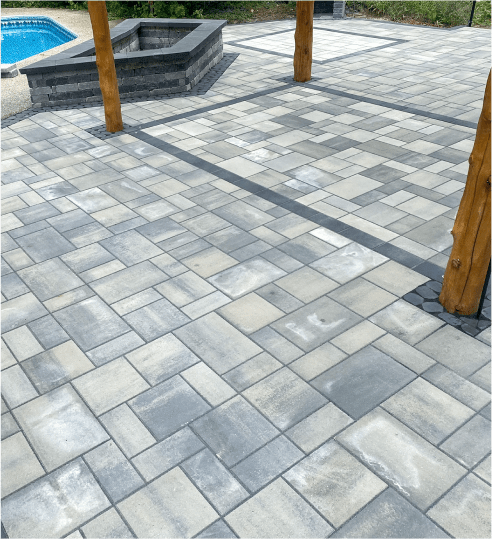 A spacious patio with gray stone tiles, wooden posts, and a nearby fire pit, adjacent to a pool and greenery. No persons visible.