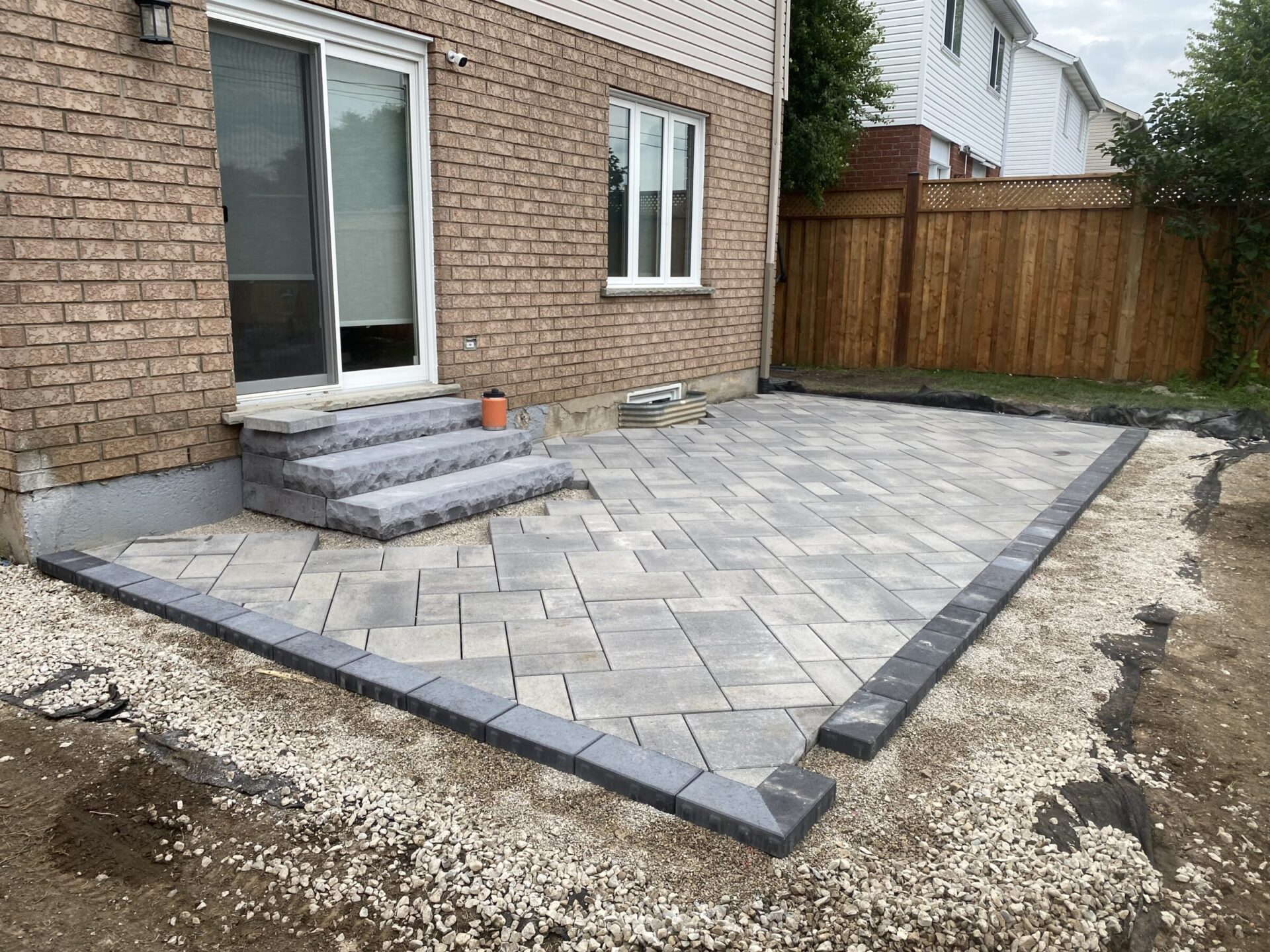 A newly paved patio with grey bricks adjacent to a brick house, surrounded by gravel and enclosed by a wooden fence.