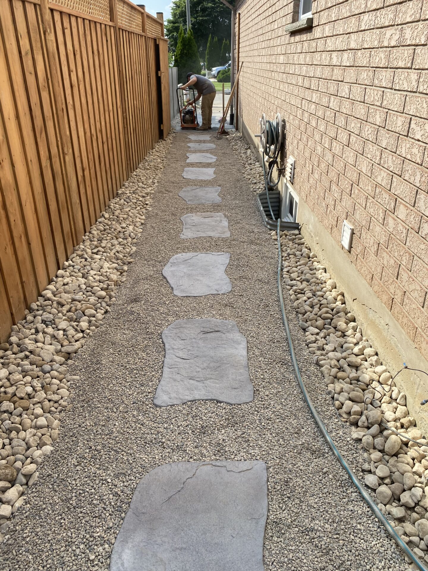 A person uses equipment to lay a stone pathway beside a brick house, bordered by pebbles and a wooden fence.