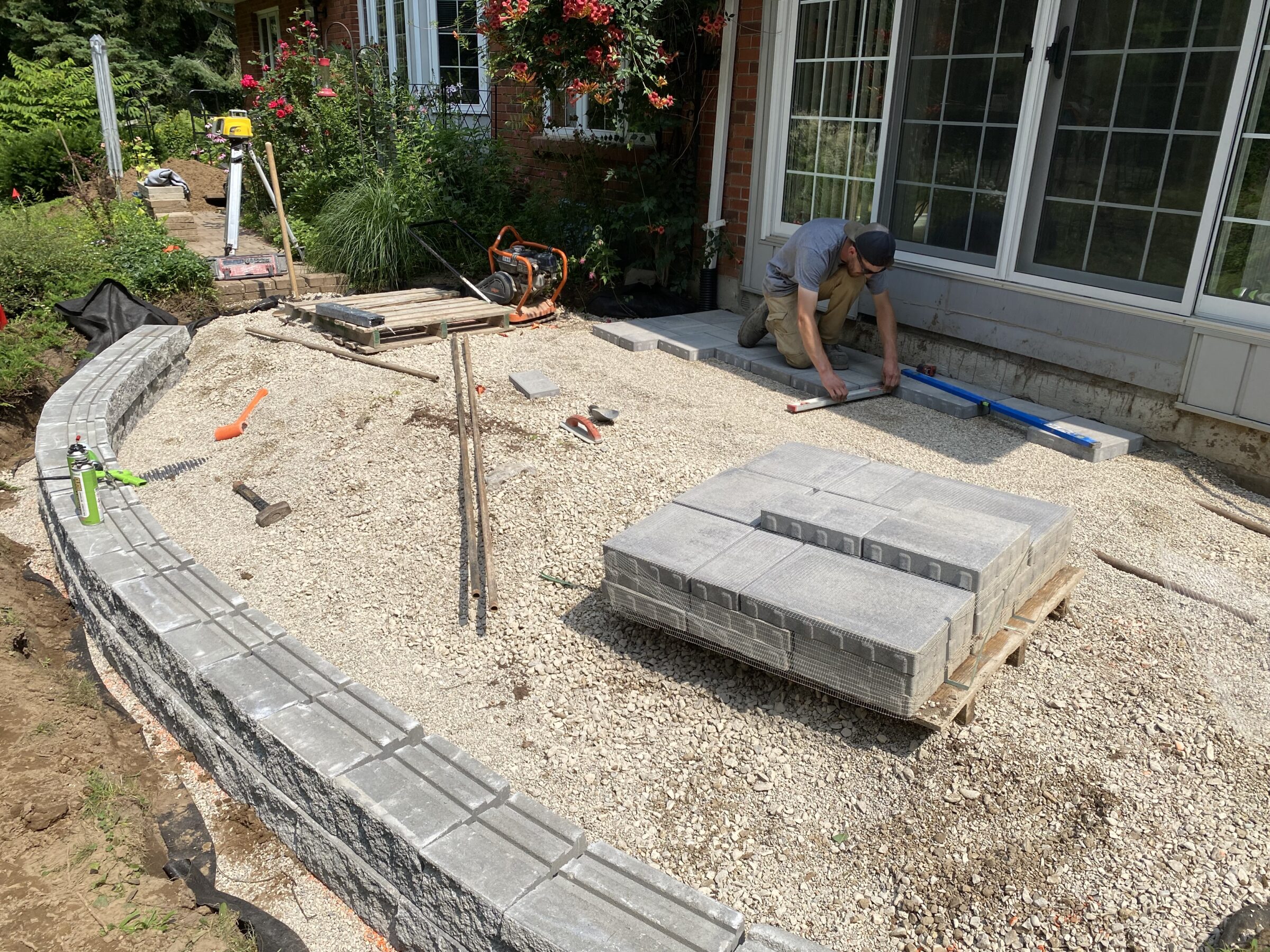 A person works on a patio construction, surrounded by tools and bricks, next to a house with a garden in the background.