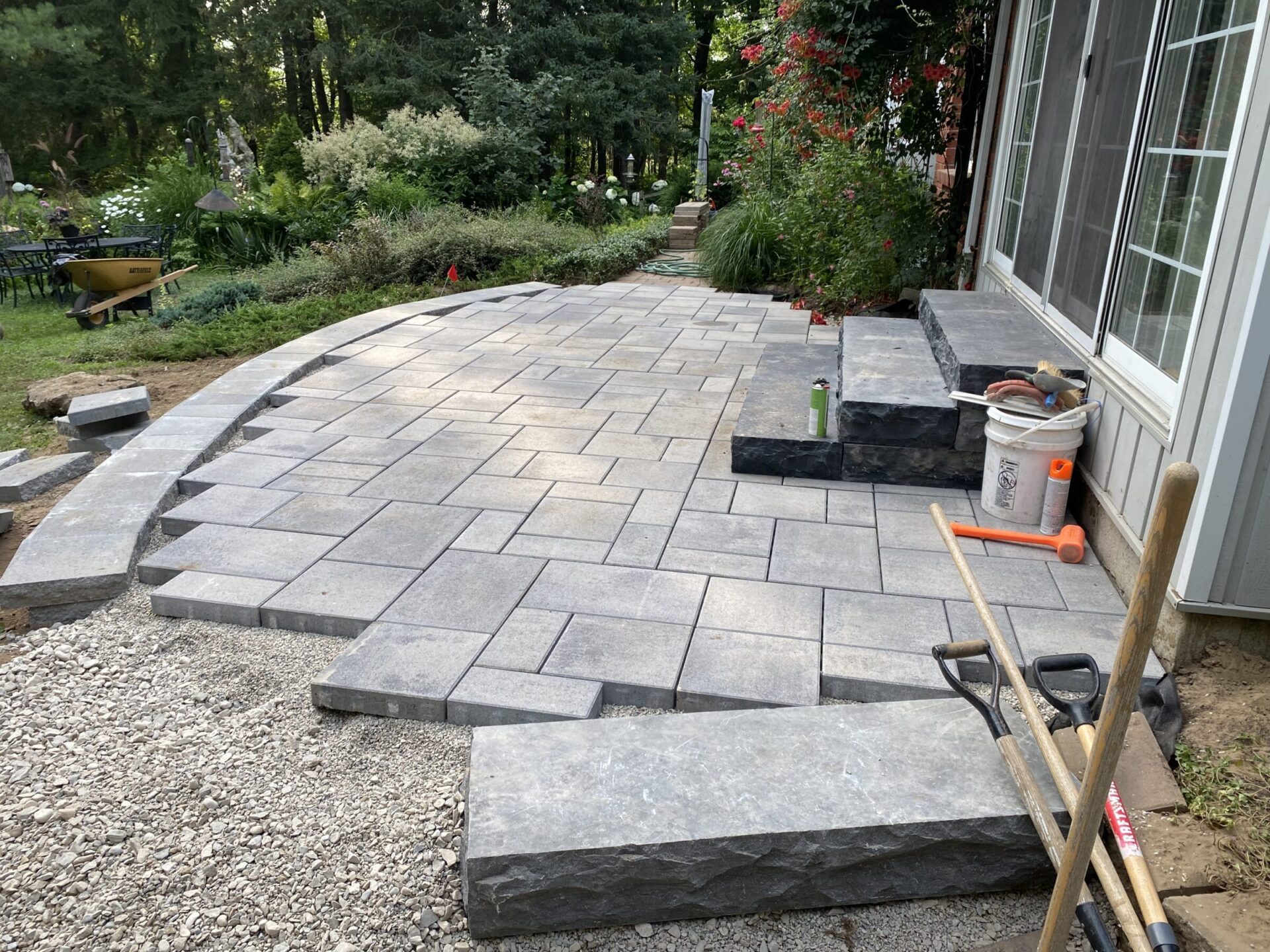 A partially completed stone patio awaits finishing touches, surrounded by lush garden greenery and tools, with steps leading to a glass door.