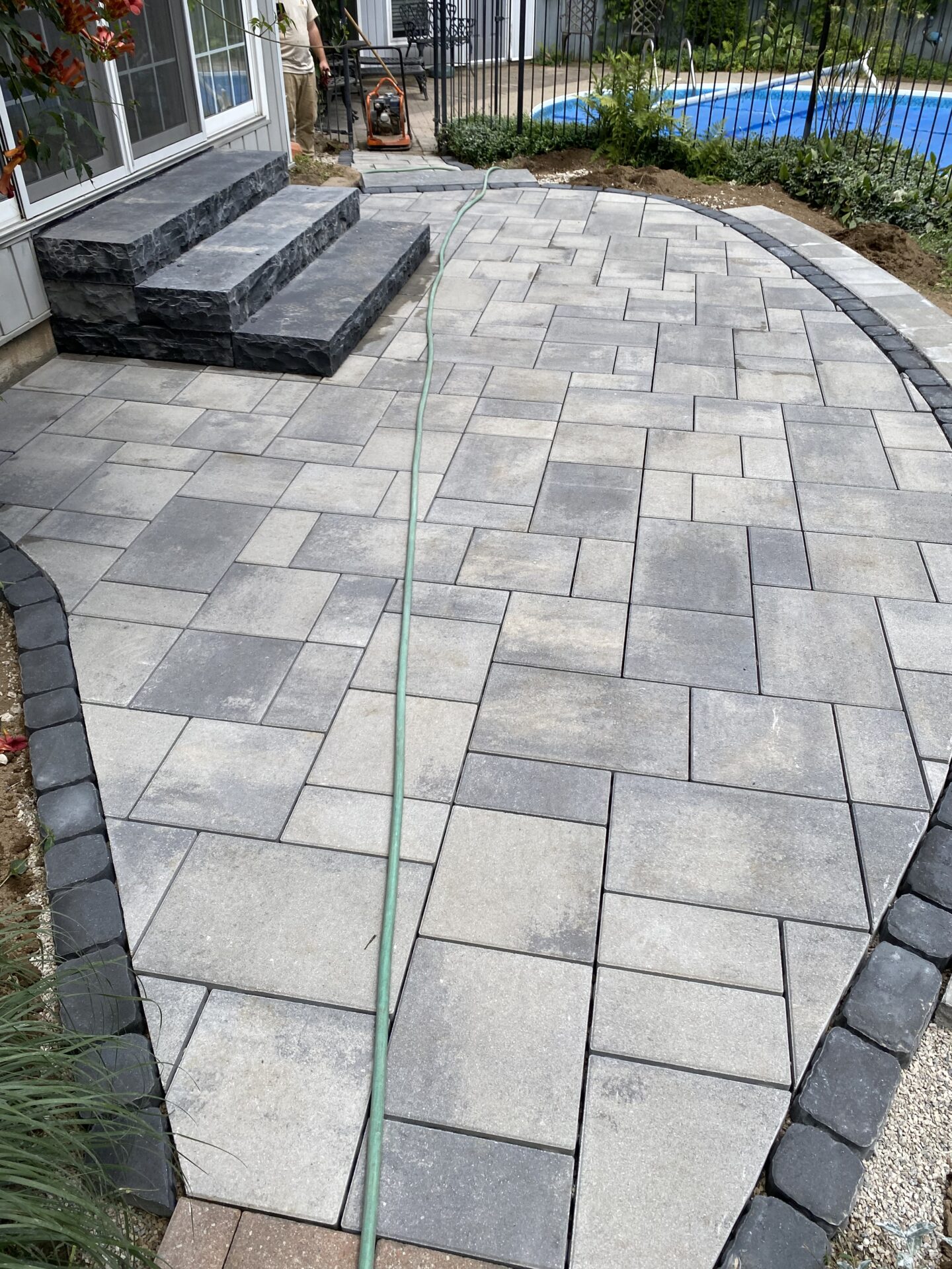 A person works on a stone patio beside a house with steps leading to a pool and garden, using a hose and equipment.