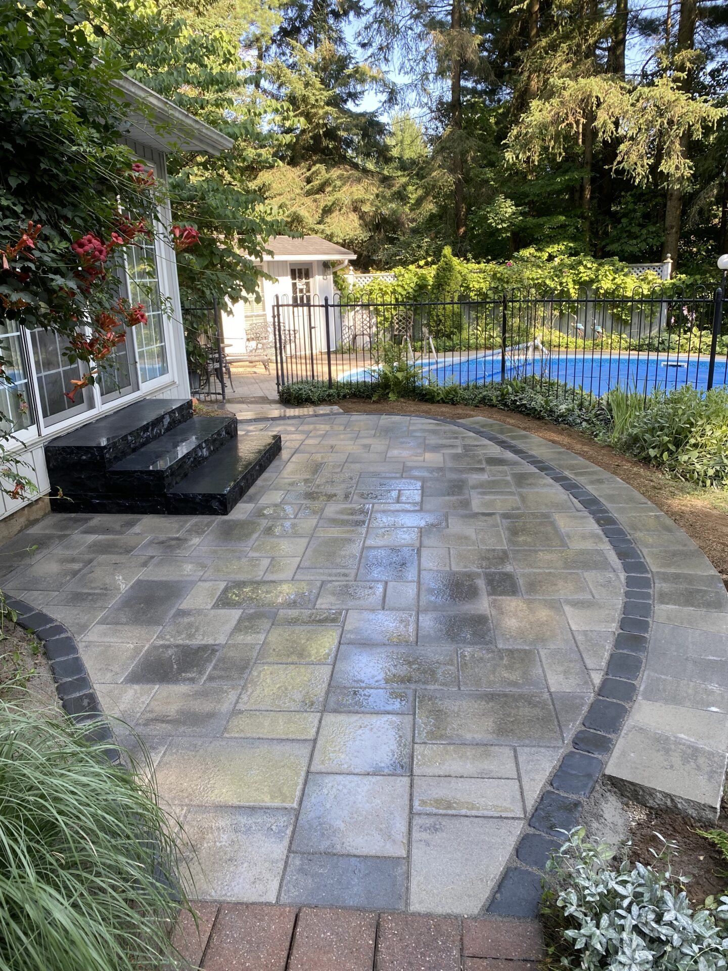 Patio with patterned tiles leads to a fenced pool area. Surrounded by lush trees, a house and steps are partially visible.