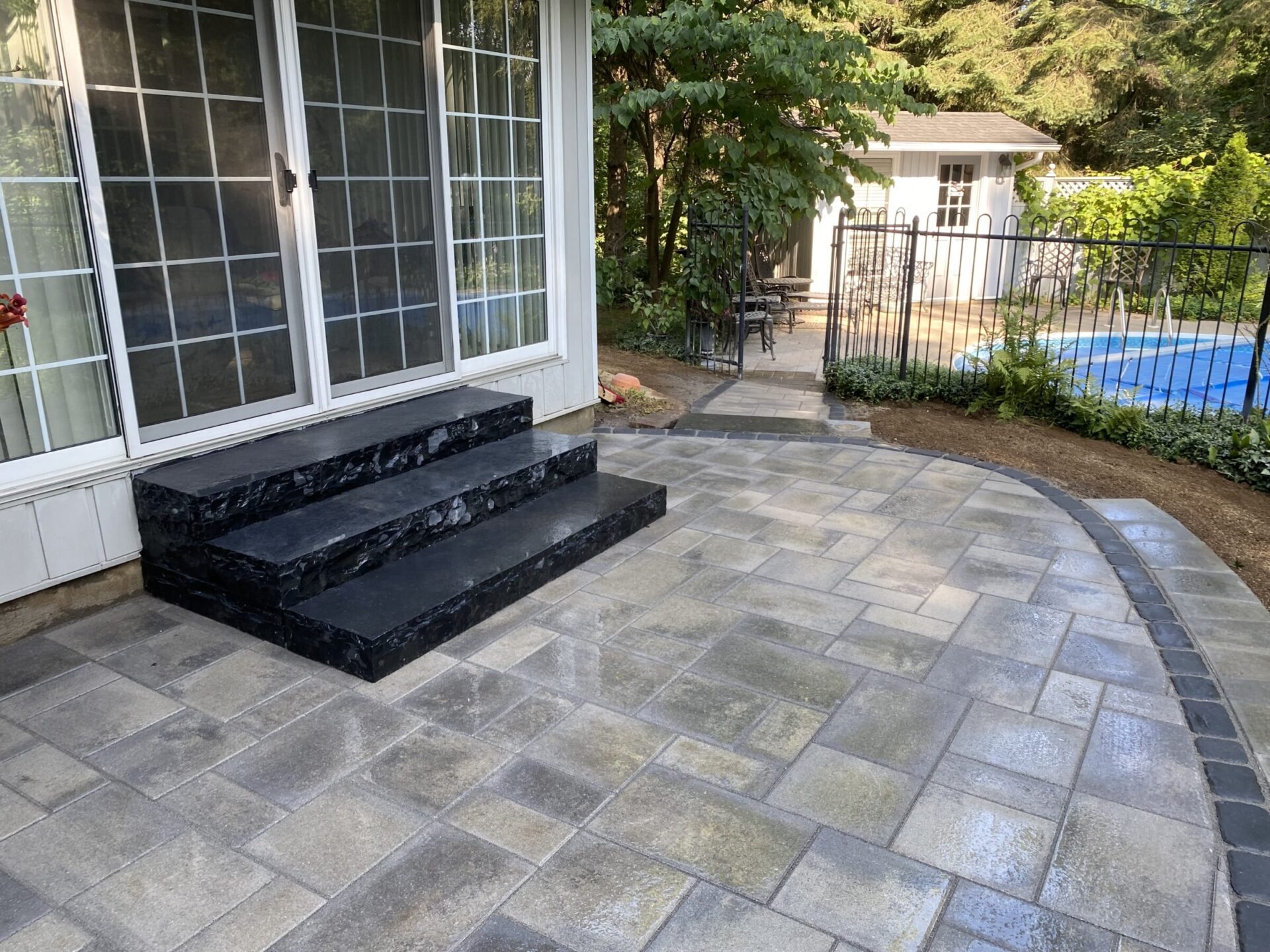 Stone patio with black steps leads to a sliding glass door. Fenced backyard features a small pool and garden shed.