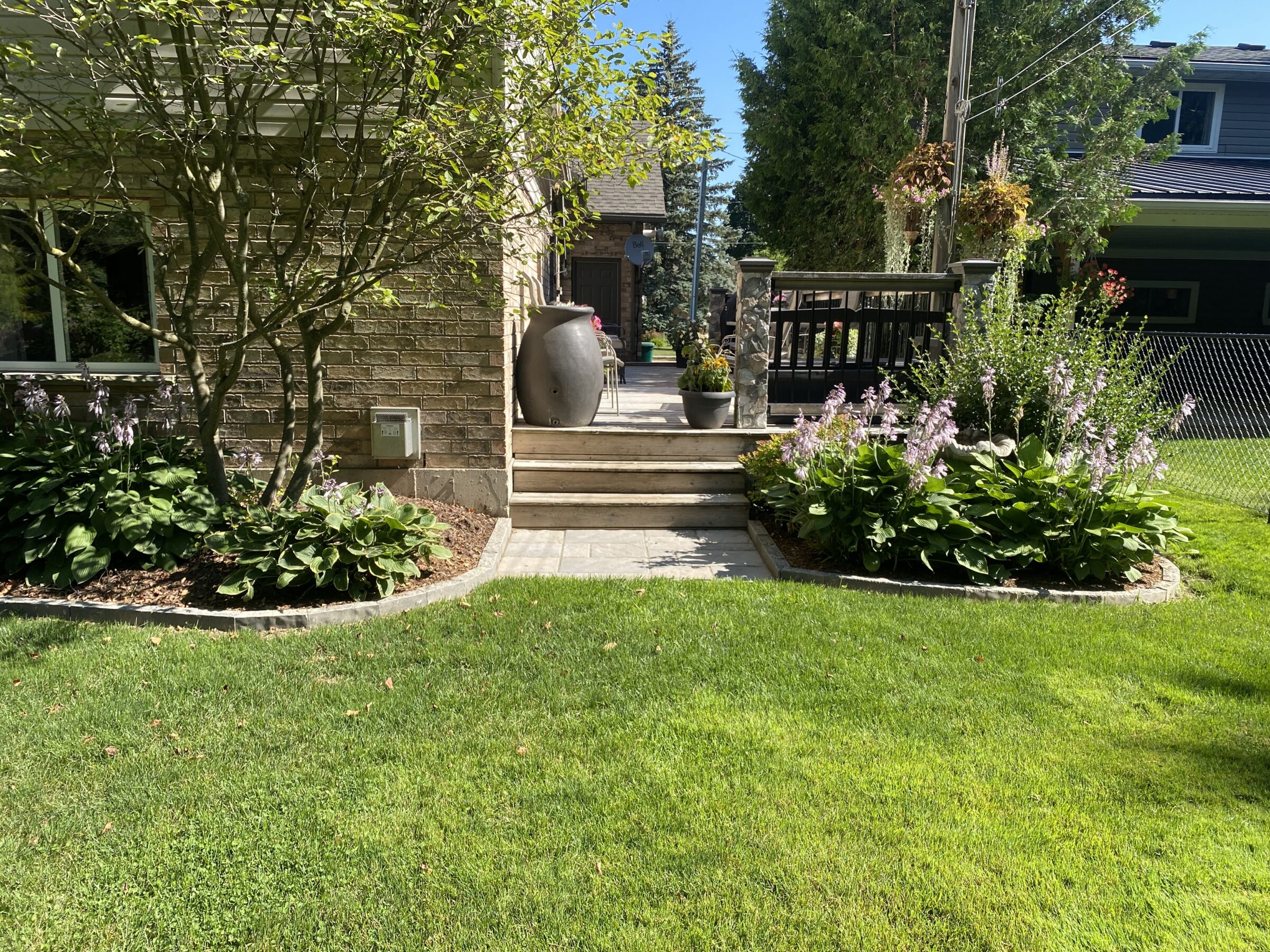 A well-maintained garden with green grass, flowering plants, and a patio with decorative pots. Brick house and wooden stairs in the background.