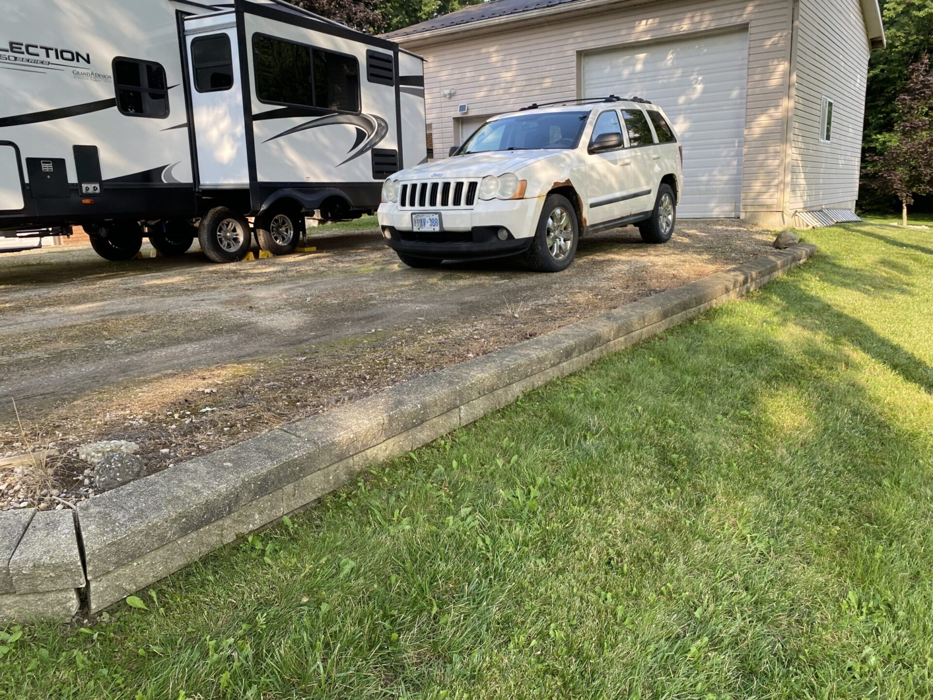 A driveway with a parked white SUV and a trailer next to a garage, surrounded by grass and trees. No people visible.