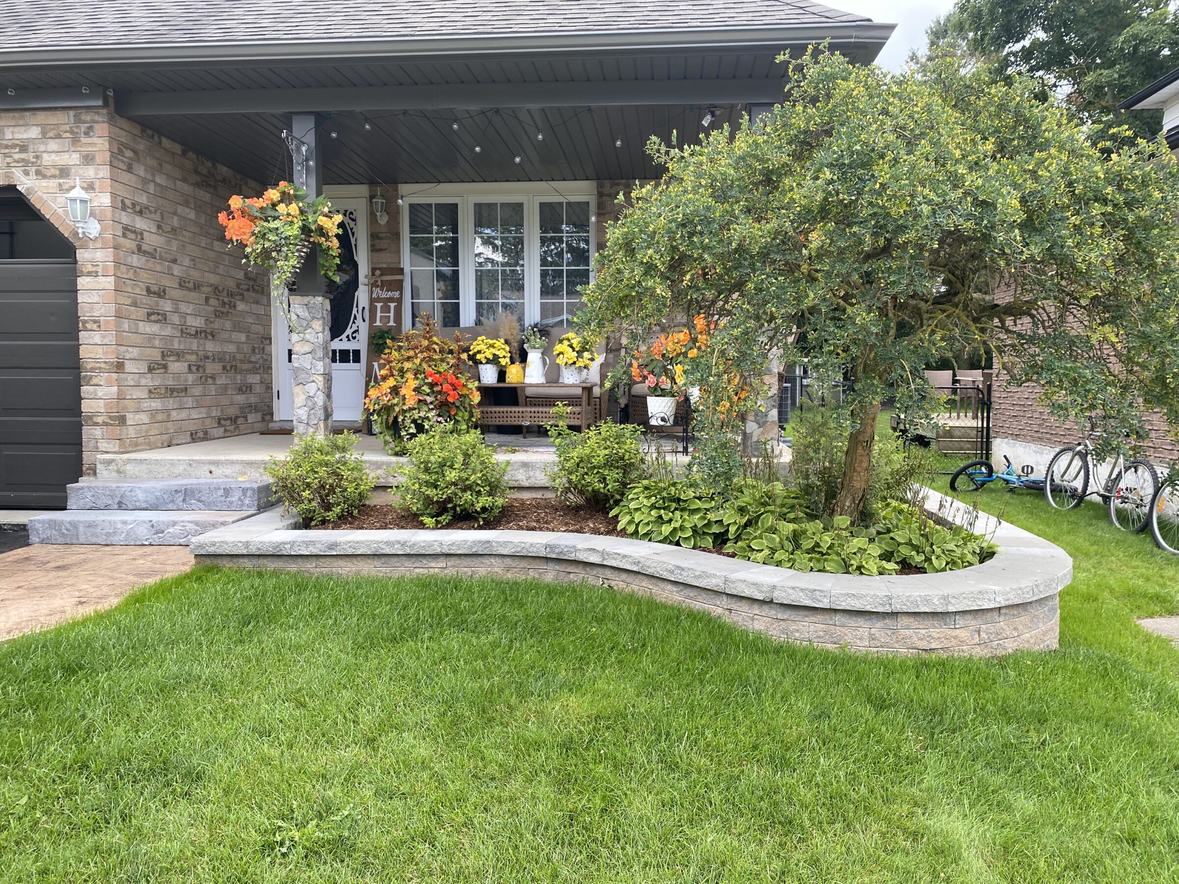 Cozy home entrance with lush greenery and colorful flowers. Bicycles rest by a brick wall, enhancing the welcoming suburban scene.