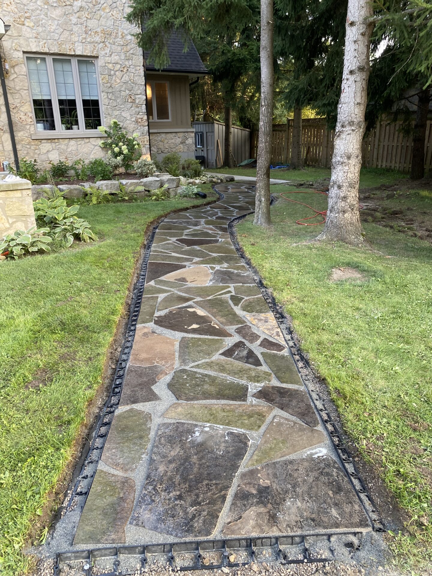 Stone path winds through garden, leading to charming stone house. Trees and lush greenery surround the area, creating a serene, natural atmosphere.