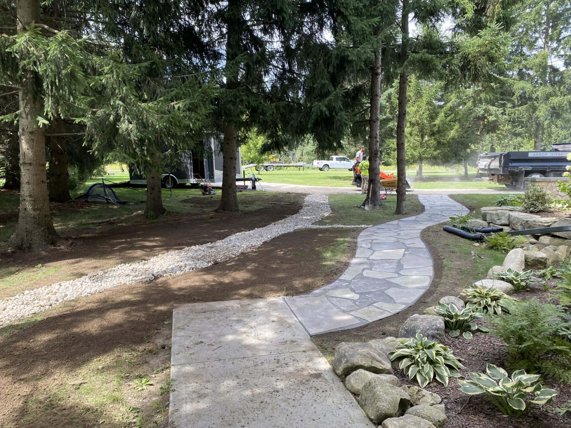 A winding stone path leads through a garden with ferns and hostas, bordered by tall trees. One person works near parked vehicles.