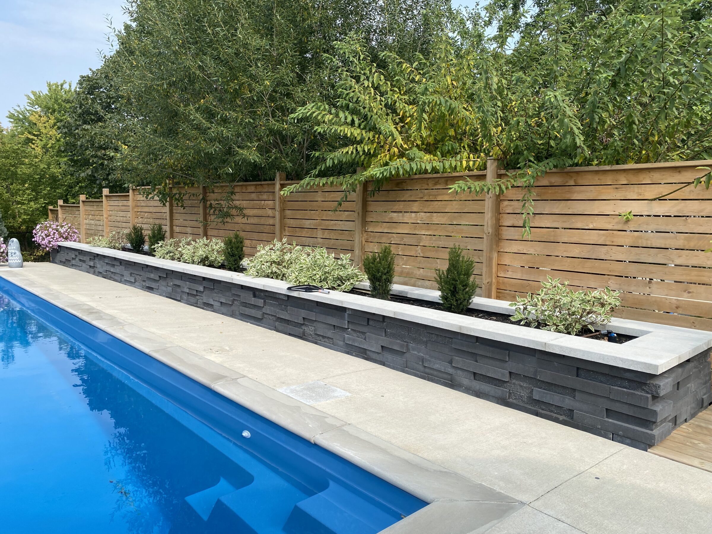 A serene backyard pool area with a sleek stone planter, vibrant greenery, wooden fence, and clear blue water reflecting the trees.