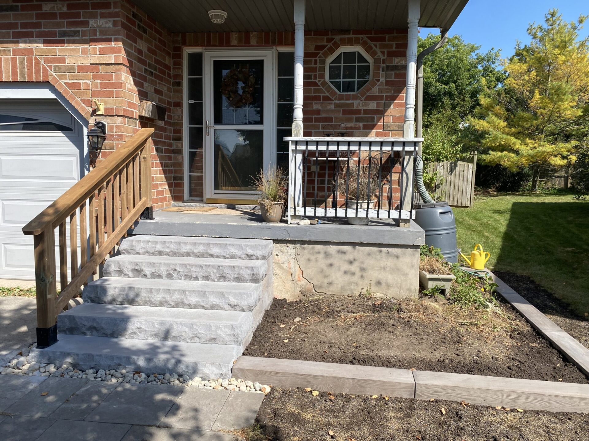 Brick house with a small porch, stone steps, and wooden railing. There’s a potted plant and a visible yellow watering can.