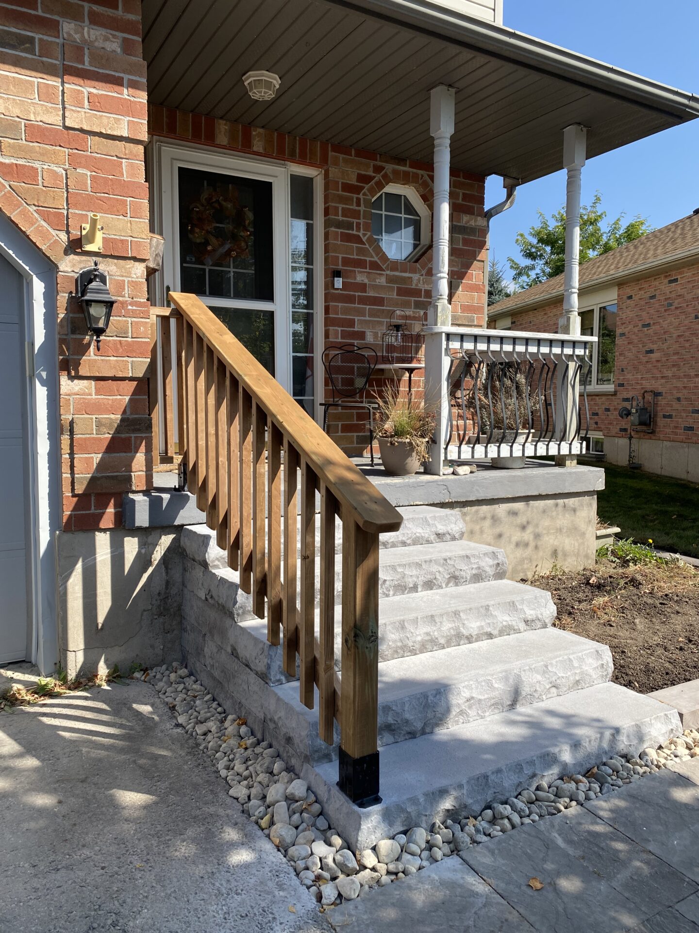 Brick house entrance with stone steps and wooden railing. Small porch with decorative chairs, potted plants, and window. Sunny day, clear blue sky.