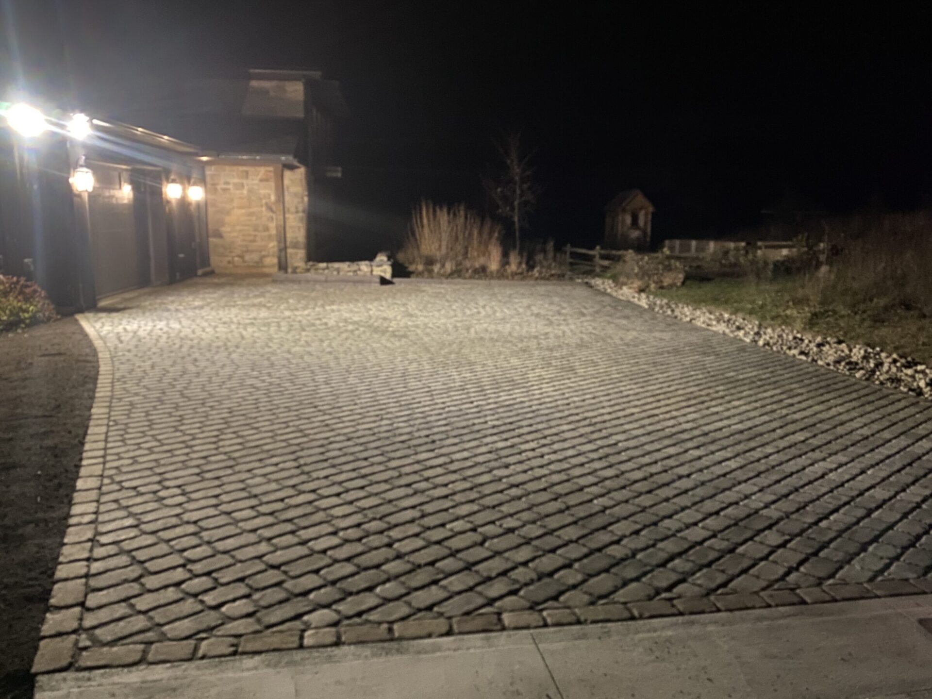 A well-lit cobblestone driveway leads to a stone building at night, surrounded by plants and a small wooden structure in the background.