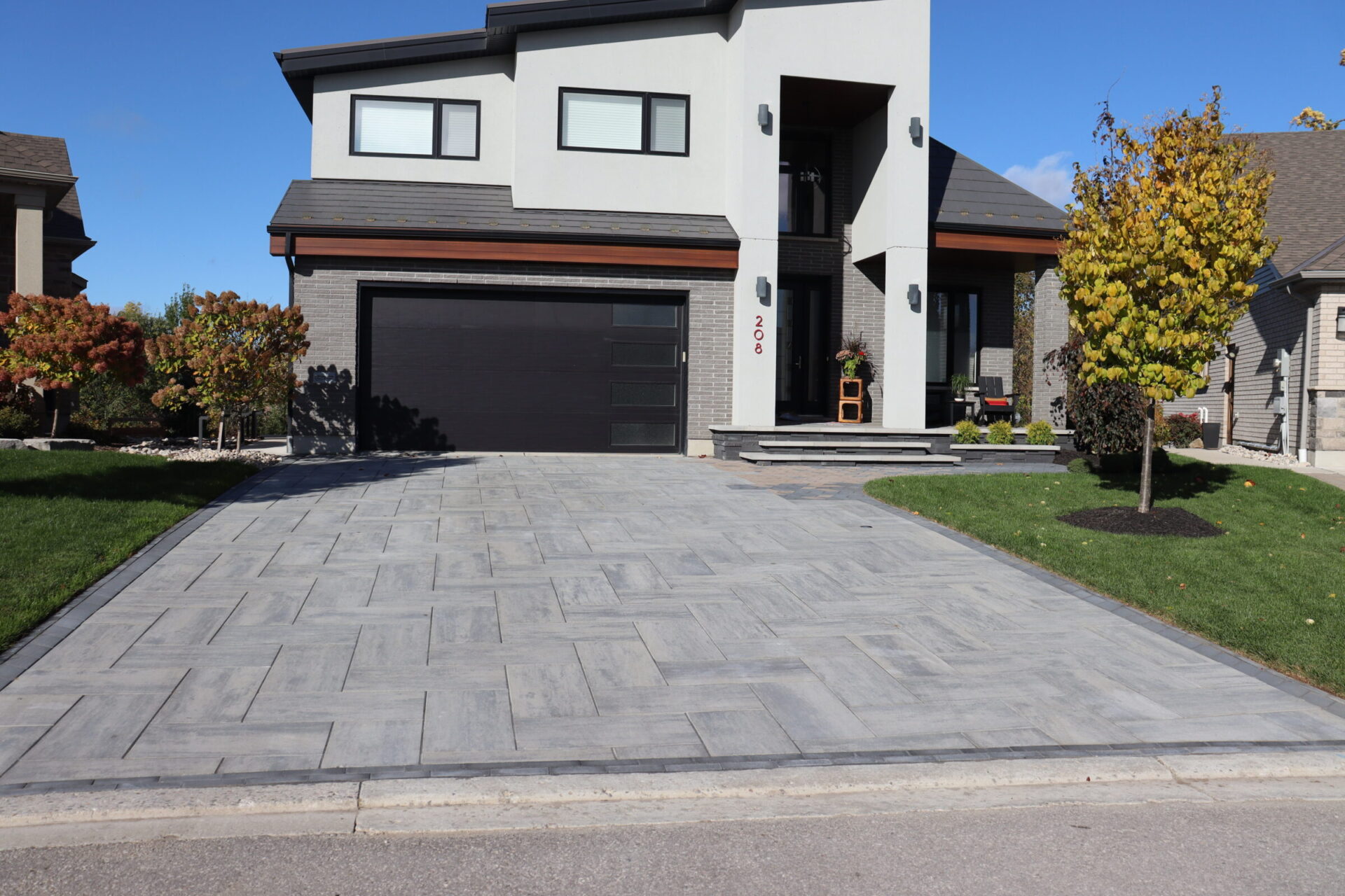 Modern two-story house with dark garage door, paved driveway, and landscaped garden featuring small trees and bushes under a clear blue sky.