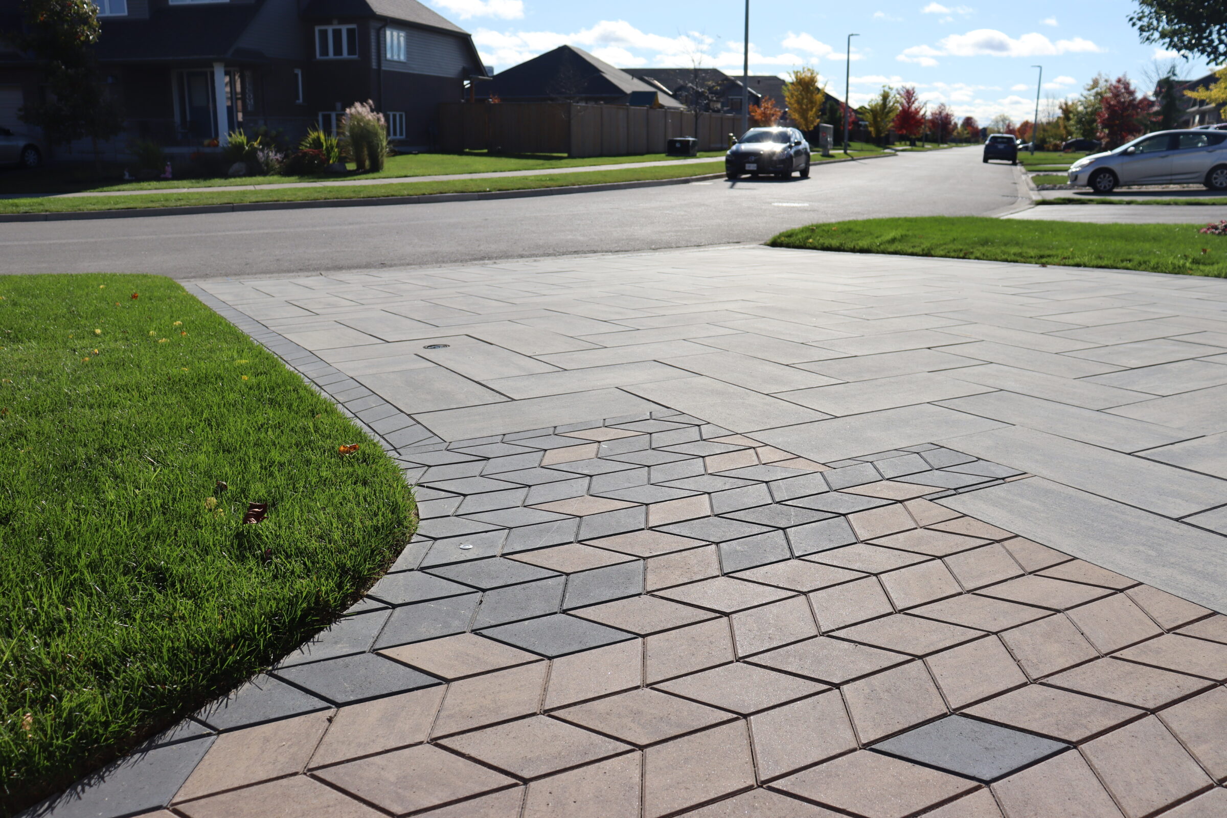 A residential street with cars parked and driving. The focus is on a detailed, geometric-patterned driveway with adjacent green grass.