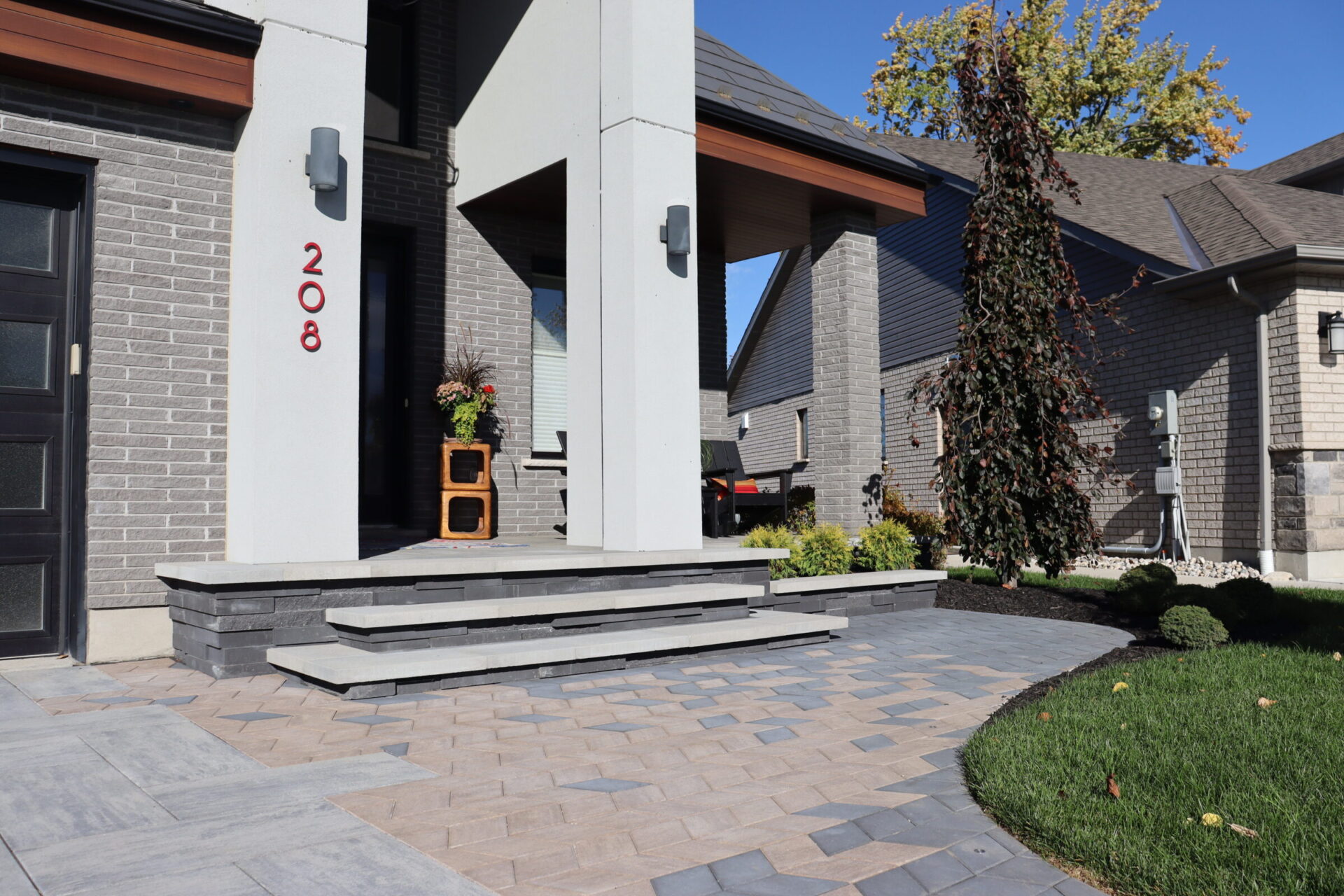 Modern house entrance with neatly landscaped yard, featuring plants and geometric paving. No people or recognizable landmarks visible. House number 208 displayed.