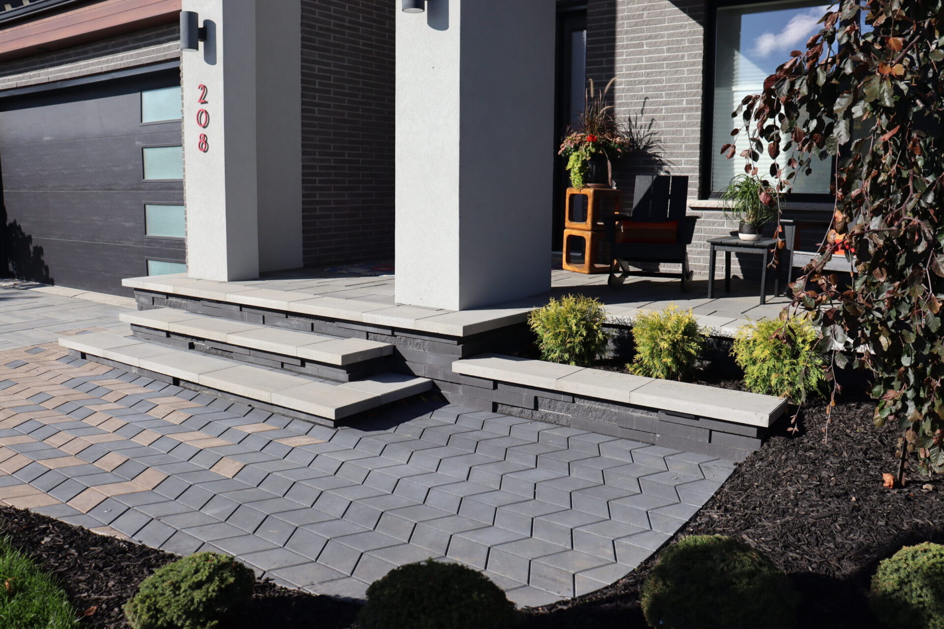 Modern house entrance with stepped porch, decorative landscaping, and brick facade. Black garage door with frosted windows and red house number 208.