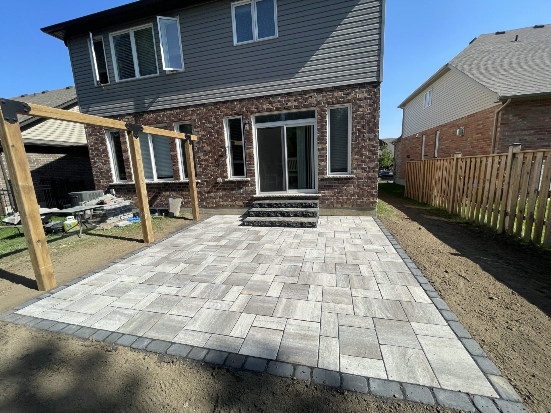 A newly constructed patio with gray stone tiles in a backyard, adjacent to a brick house, under a clear blue sky.