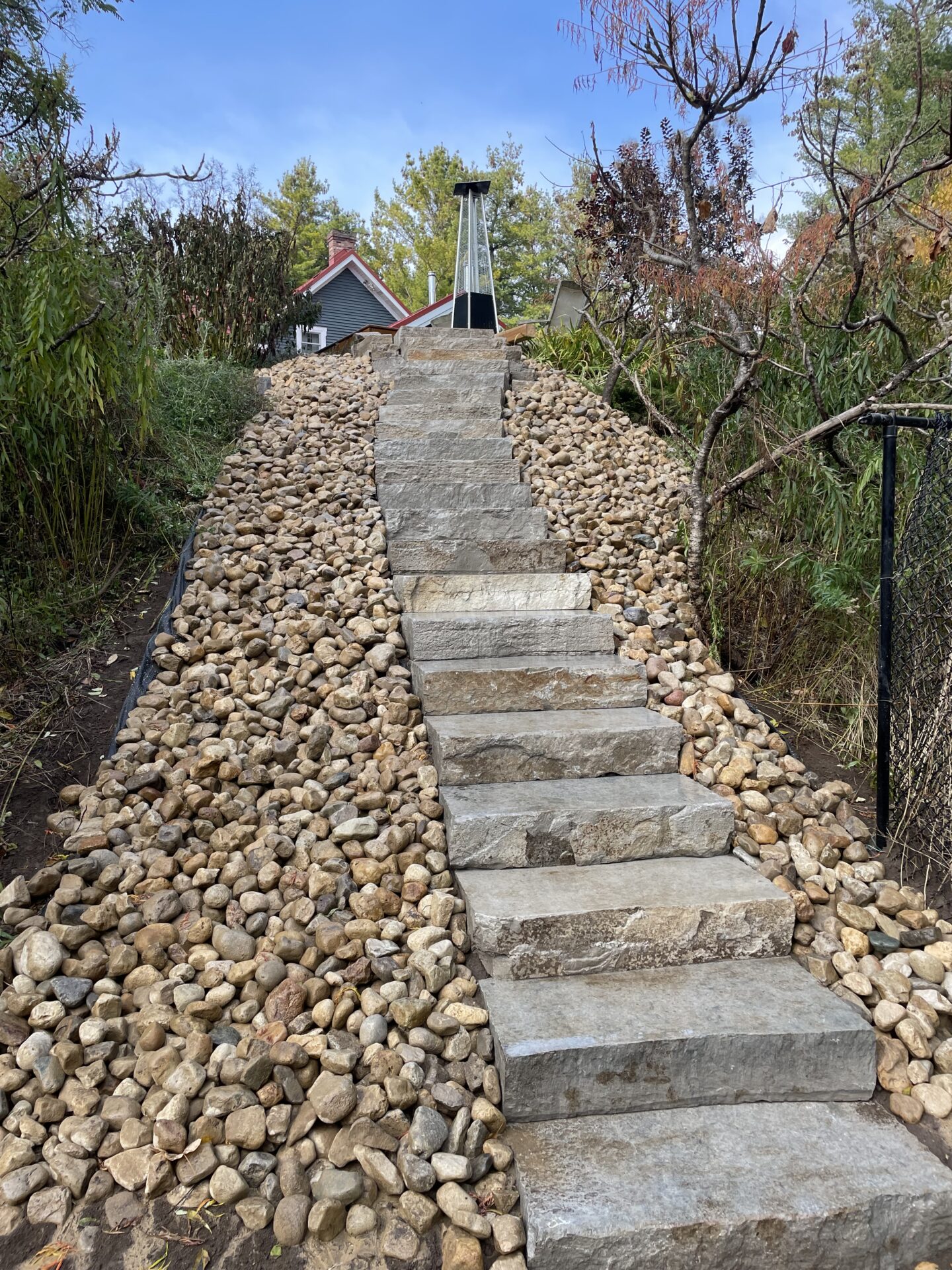 Stone steps bordered by rocks lead up to a house, surrounded by trees and a patio heater under a clear blue sky.