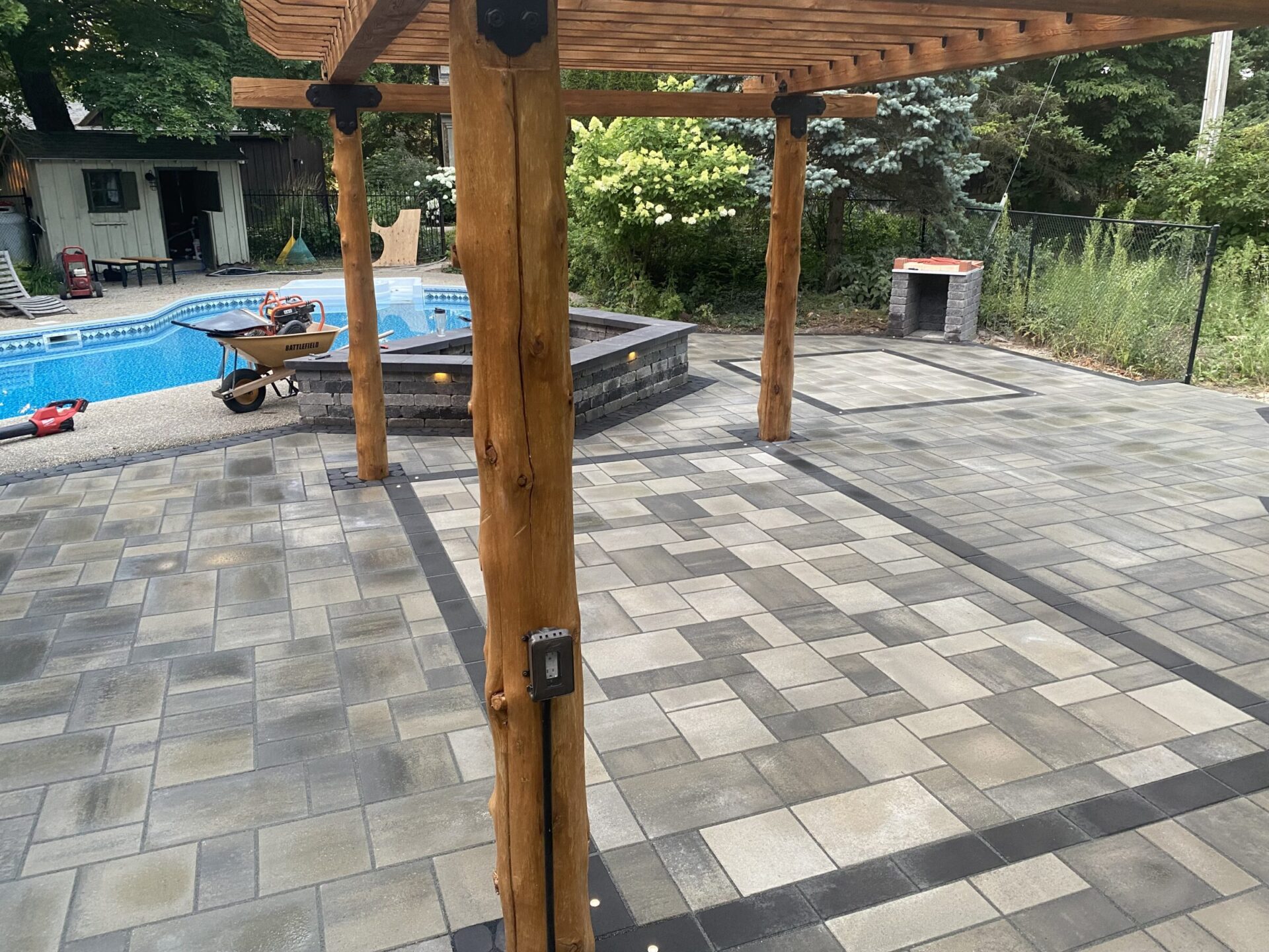 The image shows a backyard with a swimming pool, patio with pavers, wooden pergola, wheelbarrow, and surrounding greenery. No landmarks are visible.