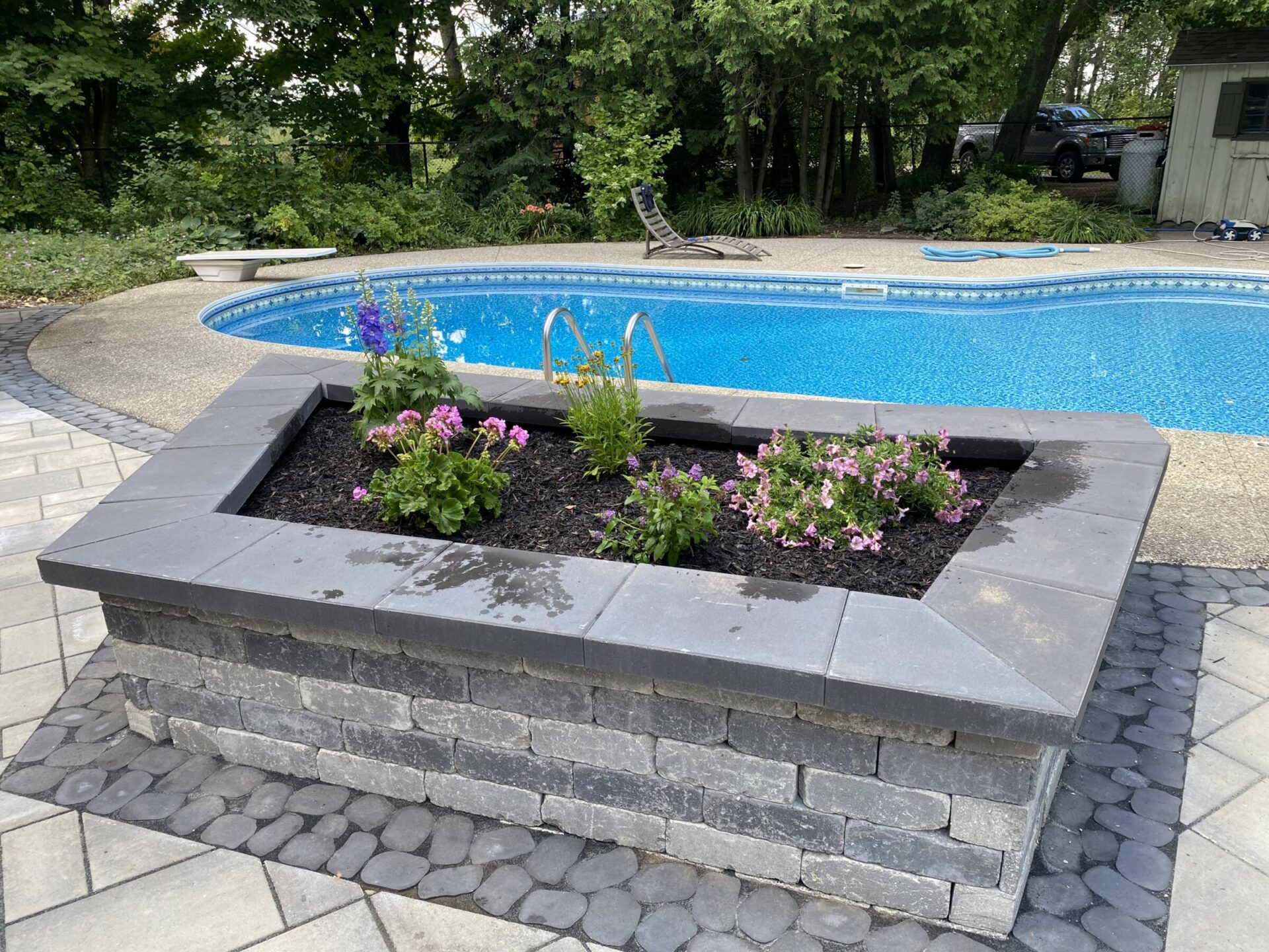 A swimming pool with a diving board, surrounded by trees and a garden bed with flowers, is adjacent to a stone patio.