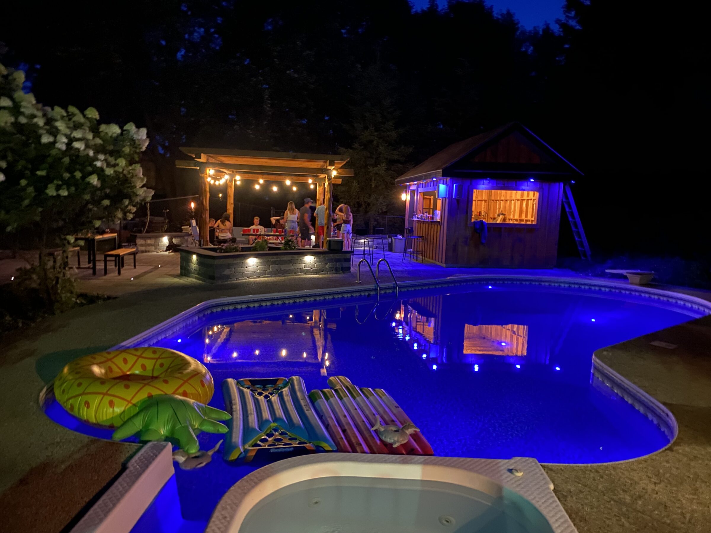 A nighttime scene with a lit pool, inflatable toys, and a wooden bar. People gather under a pergola with string lights.