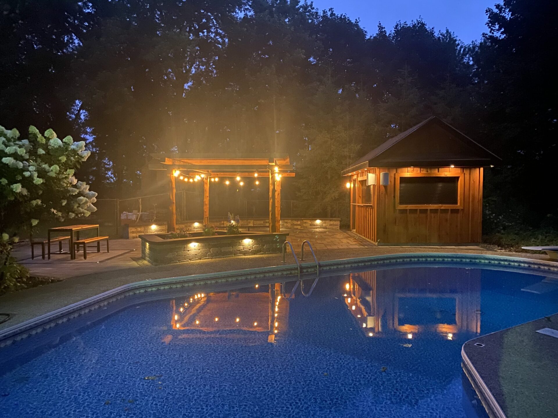 A serene evening poolside scene with glowing string lights, a wooden pavilion, and lush trees, creating a cozy and inviting atmosphere.