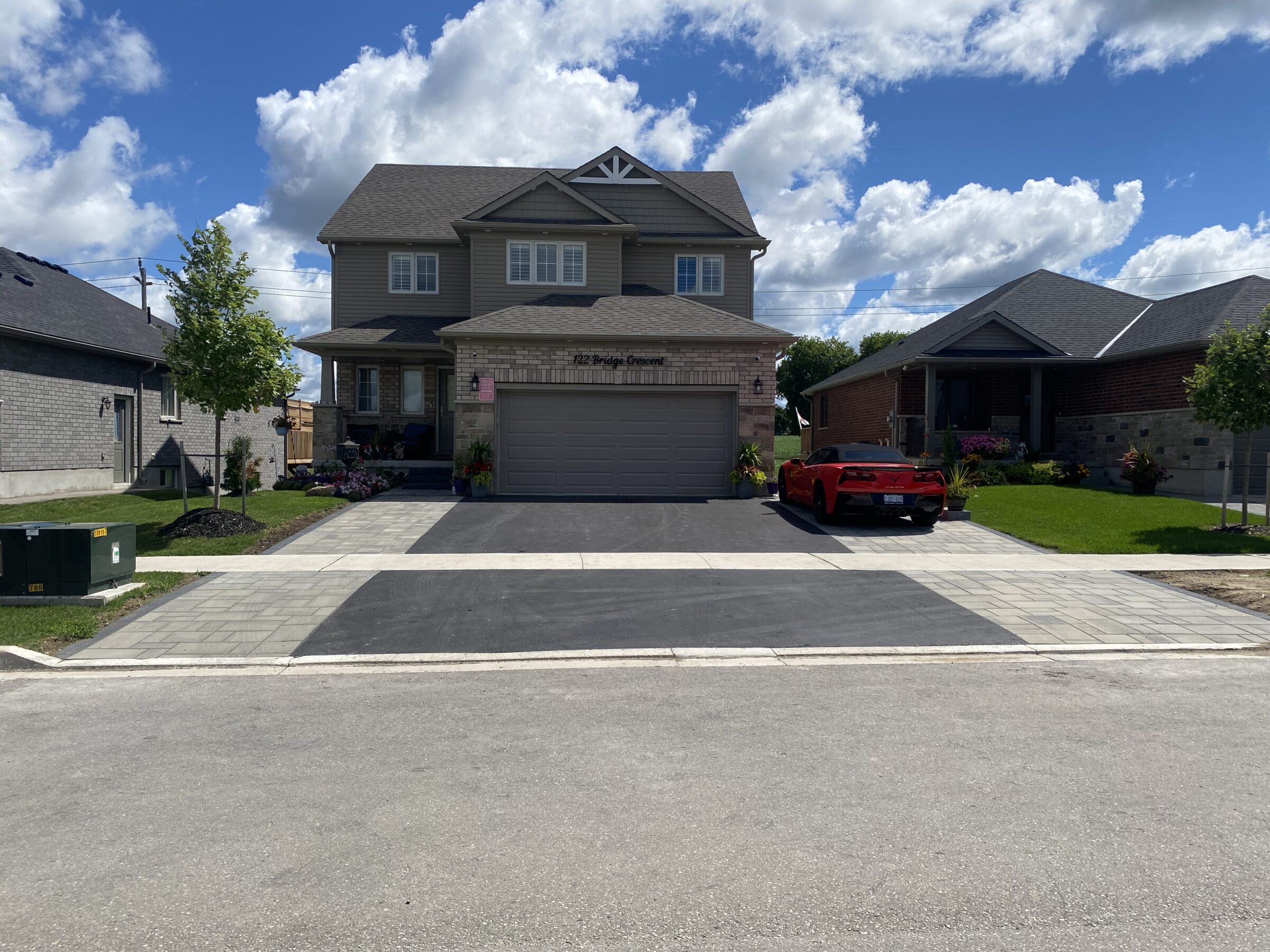 Suburban house with a red sports car in the driveway, surrounded by well-maintained lawns and clear skies with fluffy clouds.