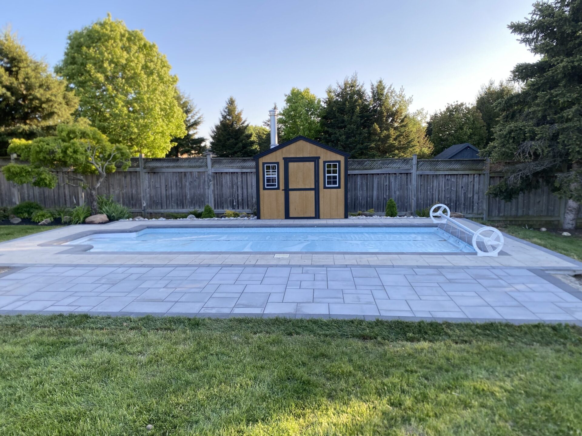 A backyard with a small wooden shed, in-ground pool with cover, surrounded by trees and a wooden fence, under a clear sky.