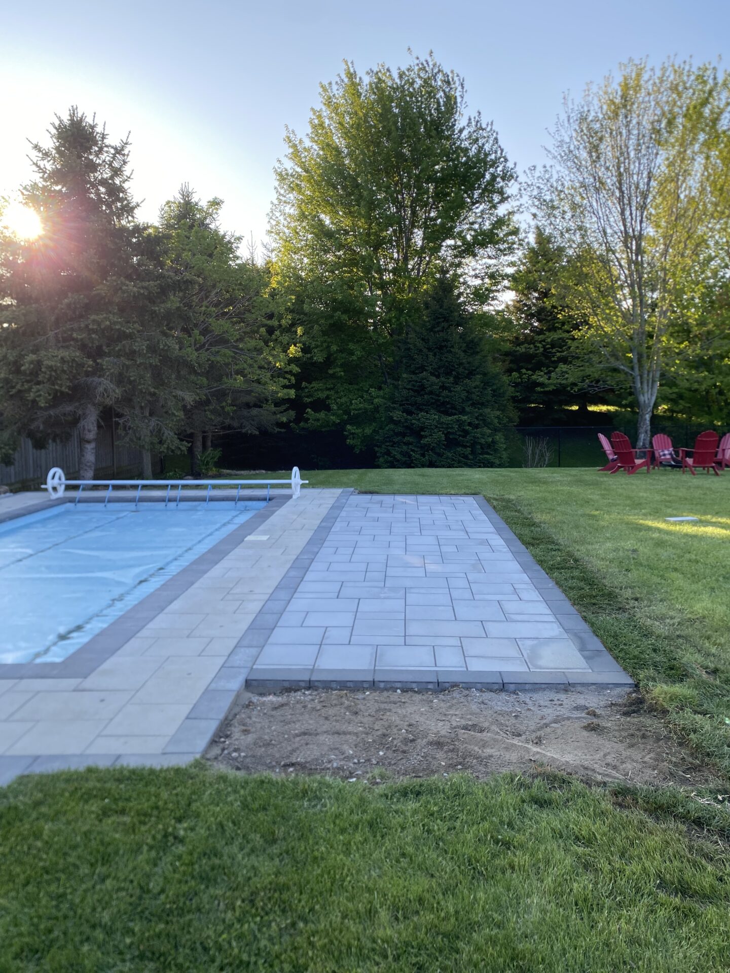 A serene backyard scene with a covered pool, patio, and red chairs. Trees and sunlight create a peaceful, natural atmosphere.