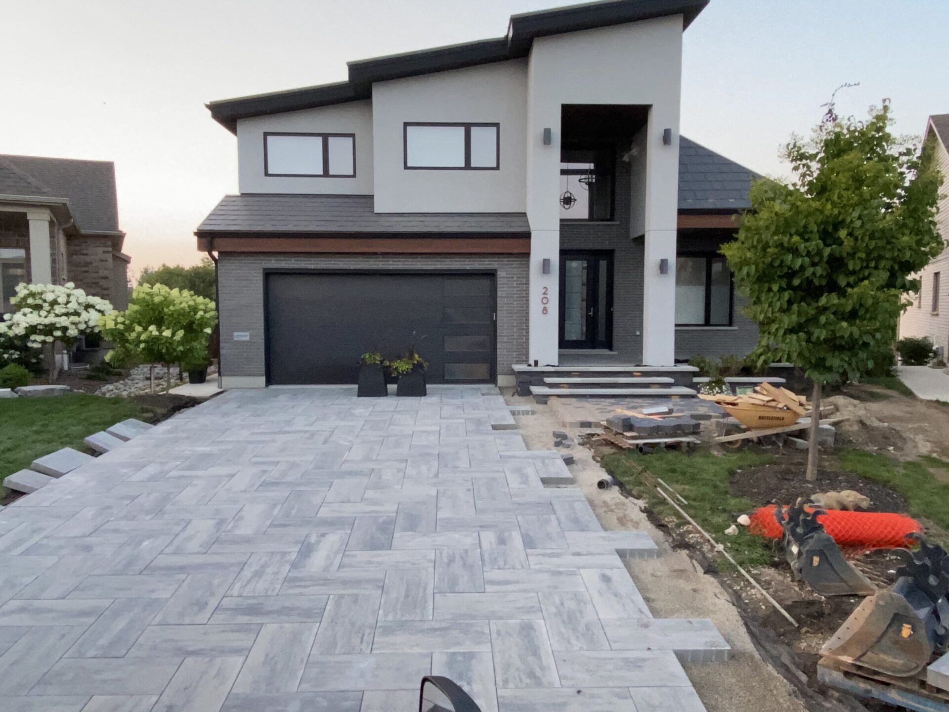 Modern house with large driveway under construction, featuring new pavers, surrounding trees, and landscaping materials. Nearby houses are visible in the neighborhood.