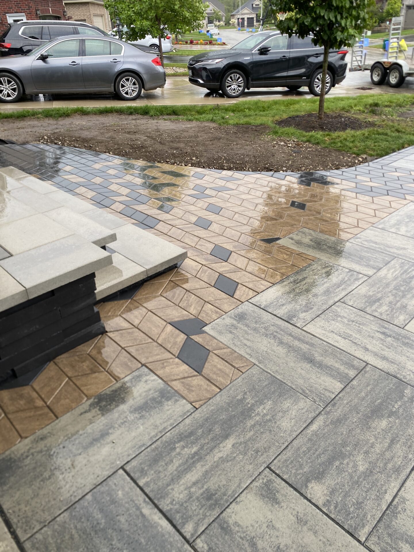 Newly laid paver patio with geometric design in a residential area. Two cars are parked on a wet street nearby.